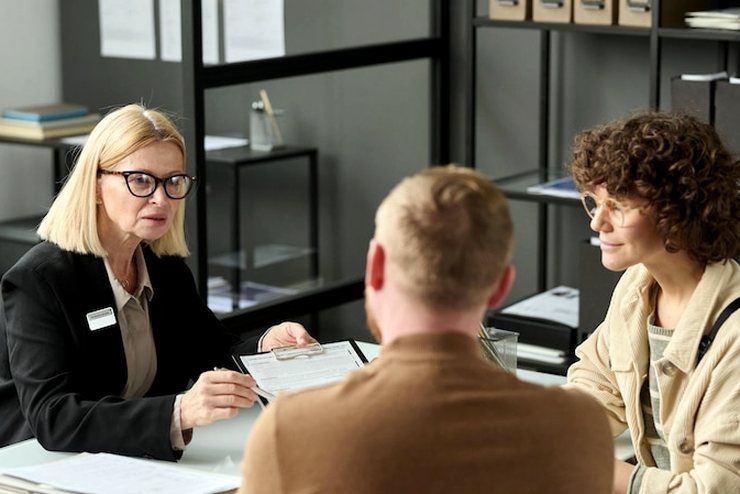 A woman with a name badge holds a clipboard while two people sit across the desk from her