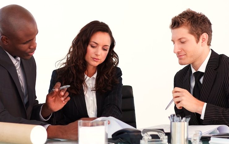 A woman and two men in dark business clothes at a table with pens, papers and a rolled document