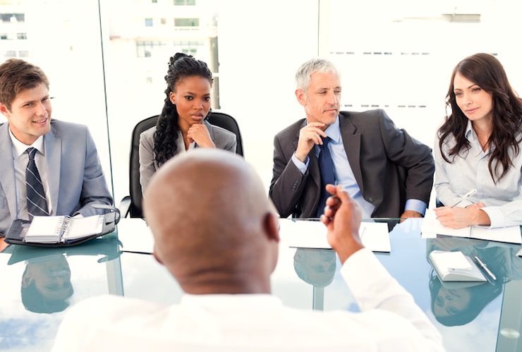 Several colleagues in business clothes around a glass conference table, one of them seen from behind in the foreground