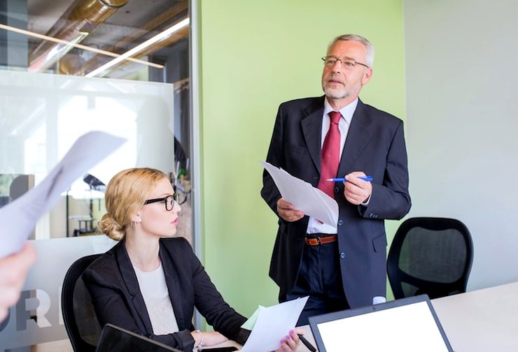 A man in a suit stands holding a sheet of paper while a woman with glasses sits at a laptop beside him