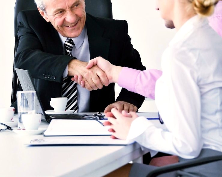An older man in a suit shakes hands with a woman across an office desk
