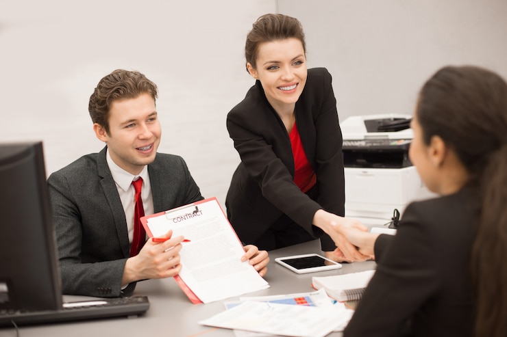 Three colleagues at an office desk, one of them handing over a printed form while another shakes hands