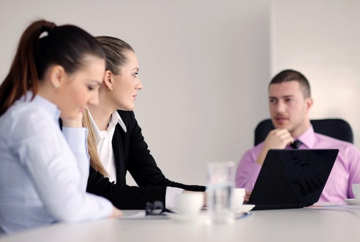Two women and a man with a laptop sit at a table with coffee cups, in conversation