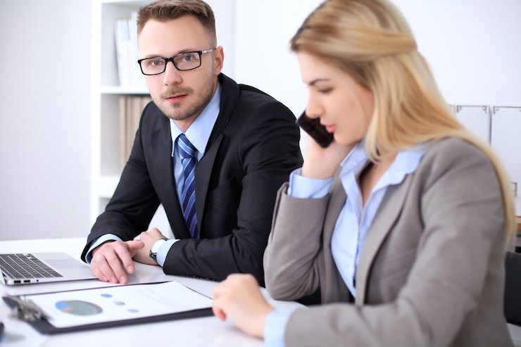 A man and a woman at a desk with a laptop and a printed chart, she is speaking on the phone